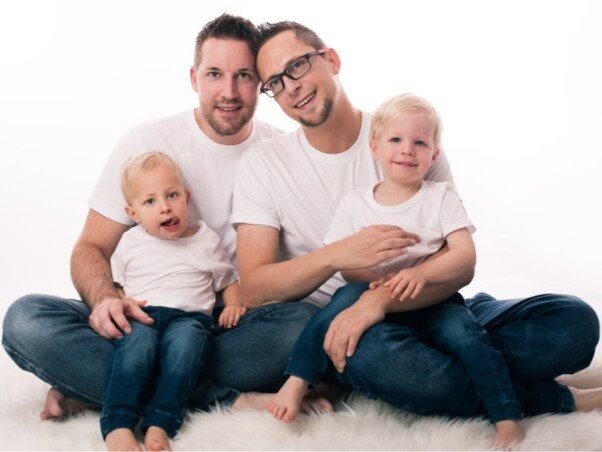 Zwei Männer mit Brillen und Bart sitzen barfuß mit ihren blonden Söhnen in weißen Shirts und Jeans auf einem flauschigen Teppich. | © privat