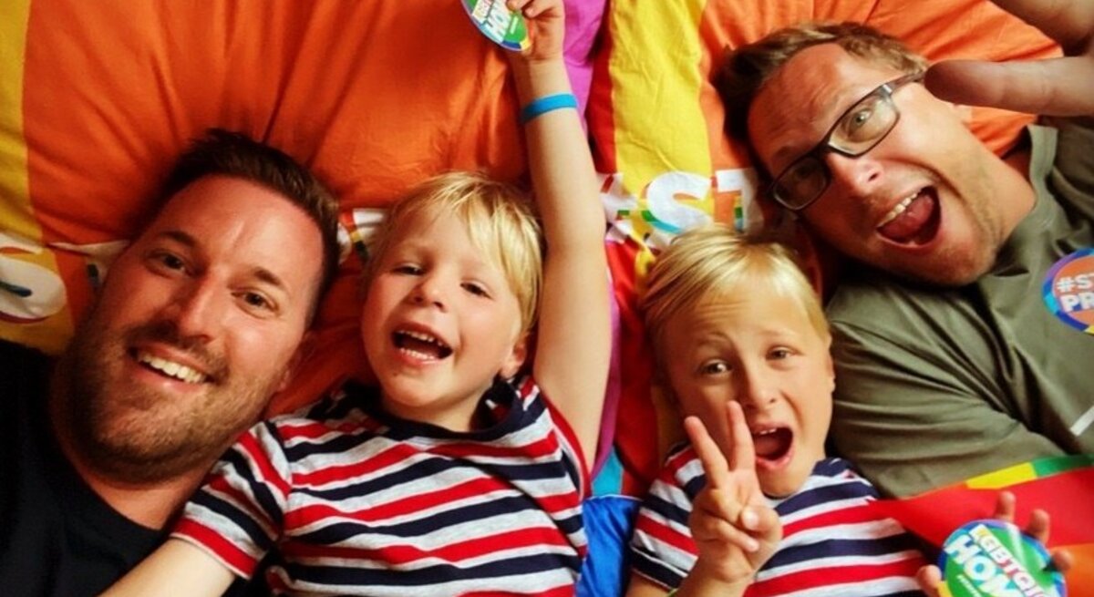 Zwei Männer und zwei blonde Jungen in gestreiften Shirts liegen lachend auf bunten Kissen, heben Arme und zeigen Friedenzeichen.
