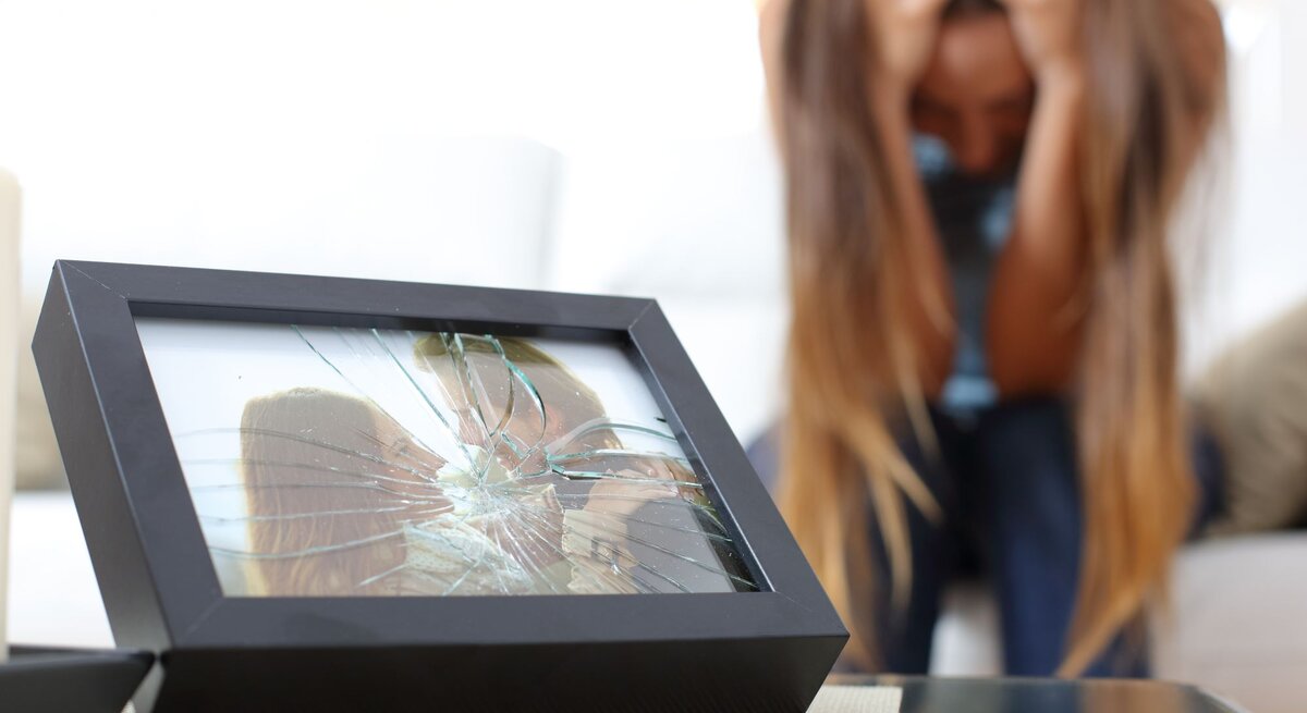 Ein zerbrochener Bilderrahmen mit einem Foto eines glücklichen Paares liegt auf einem Tisch, im Hintergrund eine verzweifelte Frau.