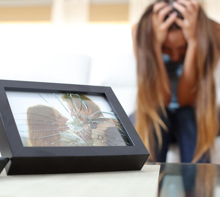 Ein zerbrochener Bilderrahmen mit einem Foto eines glücklichen Paares liegt auf einem Tisch, im Hintergrund eine verzweifelte Frau. | © Adobe Stock / Antonioguillem