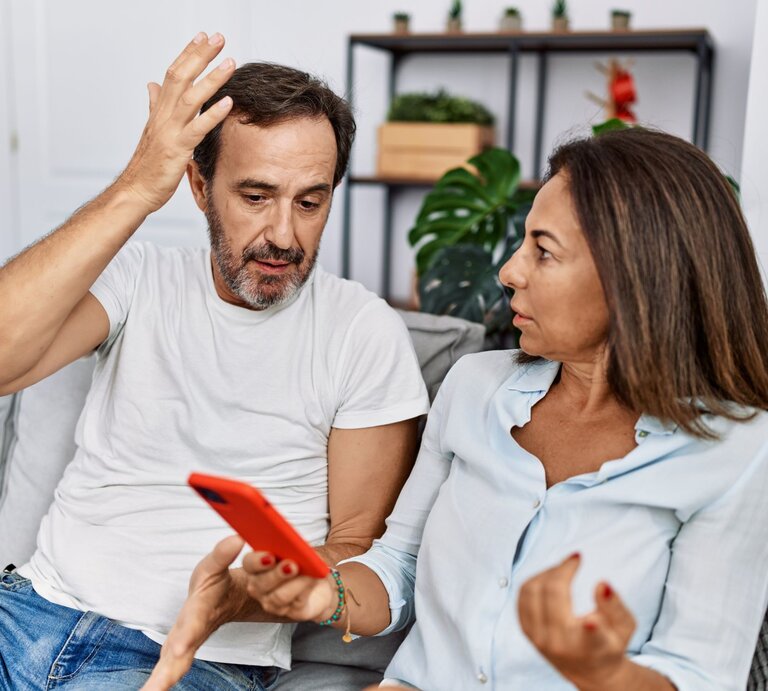Ein Mann mit Bart in weißem T-Shirt hebt frustriert die Hand, während eine Frau in hellblauer Bluse ihm mit ernster Miene ein rotes Smartphone zeigt. | © Adobe Stock / Krakenimages.com