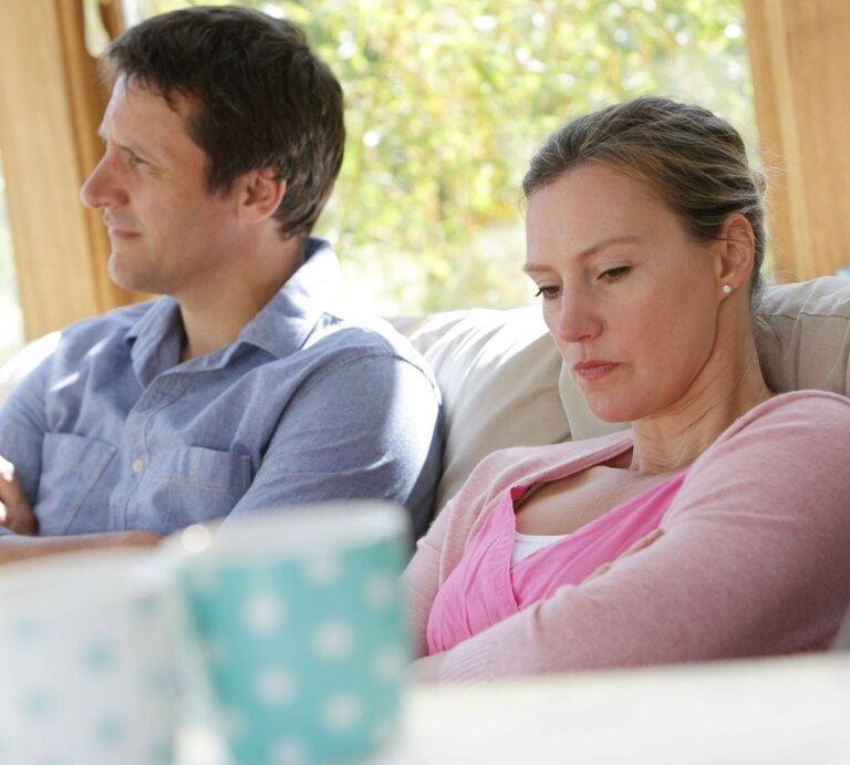 Ein Mann in blauem Hemd und eine Frau in rosa Oberteil sitzen mit verschränkten Armen schweigend auf einem Sofa und blicken angespannt weg. | © Getty Images /Peter Cade