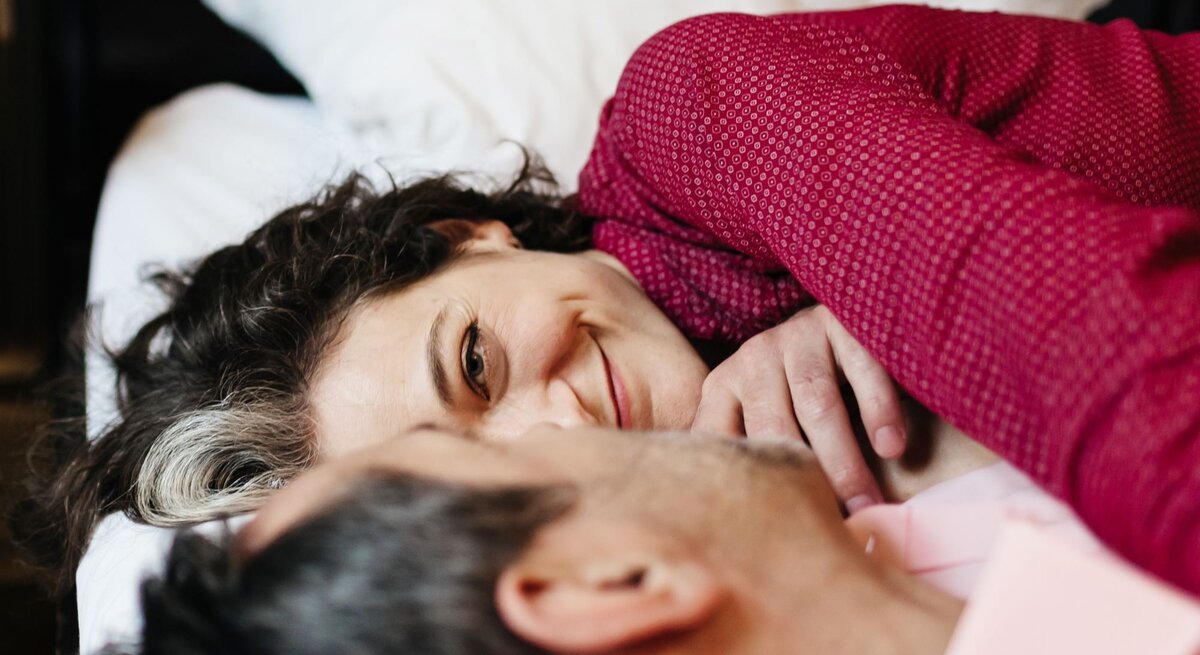 Eine dunkelhaarige Frau mit grauer Strähne lächelt zärtlich und liegt eng umschlungen mit einem Mann in rosafarbenem Hemd im Bett.