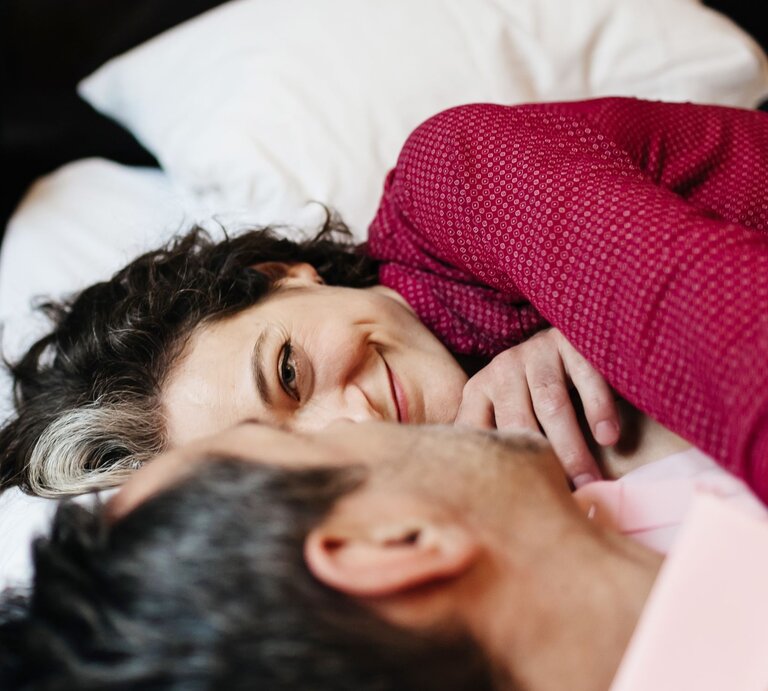 Eine dunkelhaarige Frau mit grauer Strähne lächelt zärtlich und liegt eng umschlungen mit einem Mann in rosafarbenem Hemd im Bett. | © Getty Images / Hinterhaus Productions