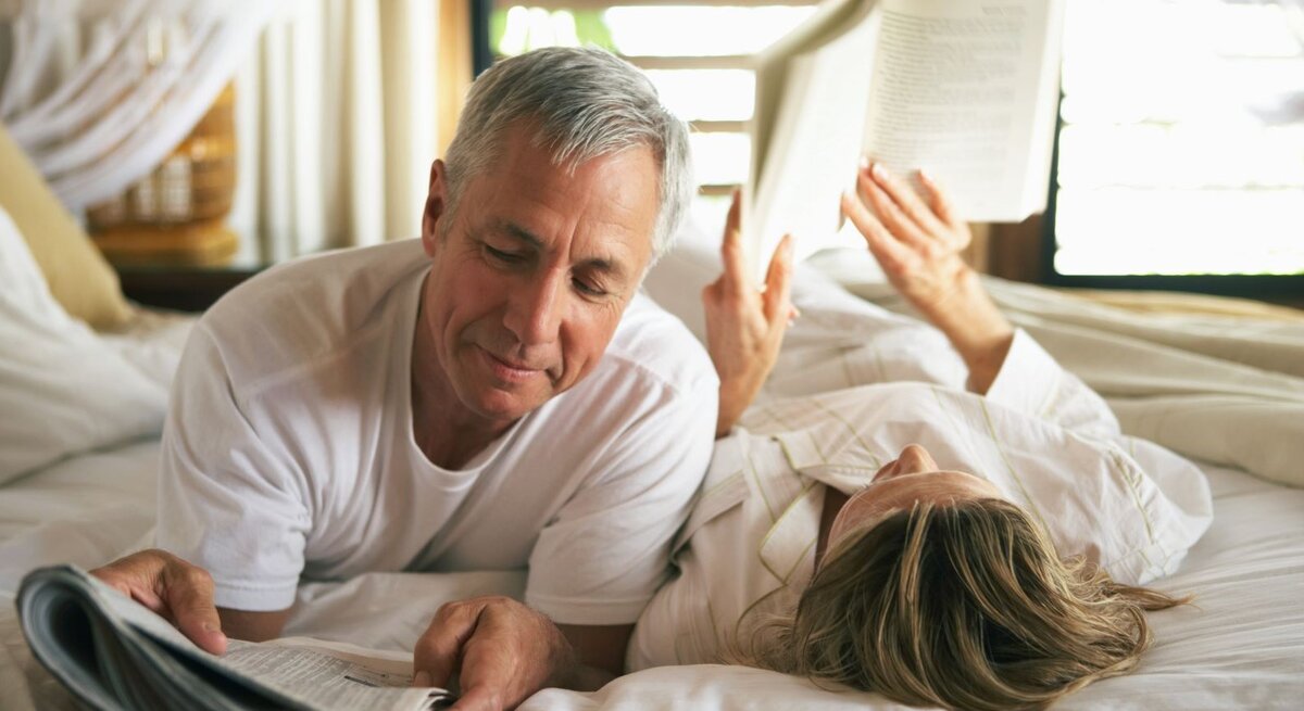 Reifes Paar liegt gemeinsam auf dem Bett, beide haben etwas zu lesen in den Händen, sie wirken glücklich und vertraut.