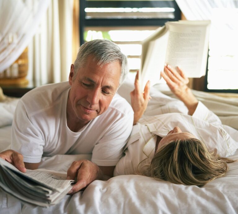 Reifes Paar liegt gemeinsam auf dem Bett, beide haben etwas zu lesen in den Händen, sie wirken glücklich und vertraut. | © Getty Images / Kraig Scarbinsky