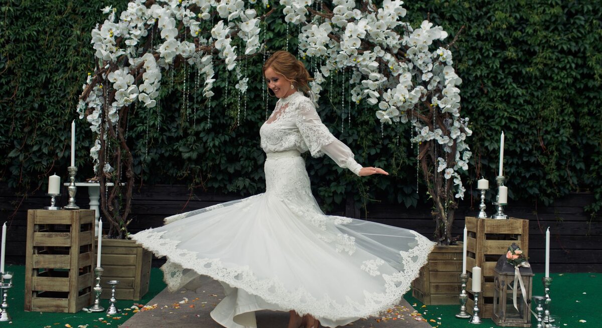 Eine lächelnde Braut mit hochgestecktem Haar dreht sich in einem weißen Spitzenkleid vor einem blumengeschmückten Hochzeitsbogen.