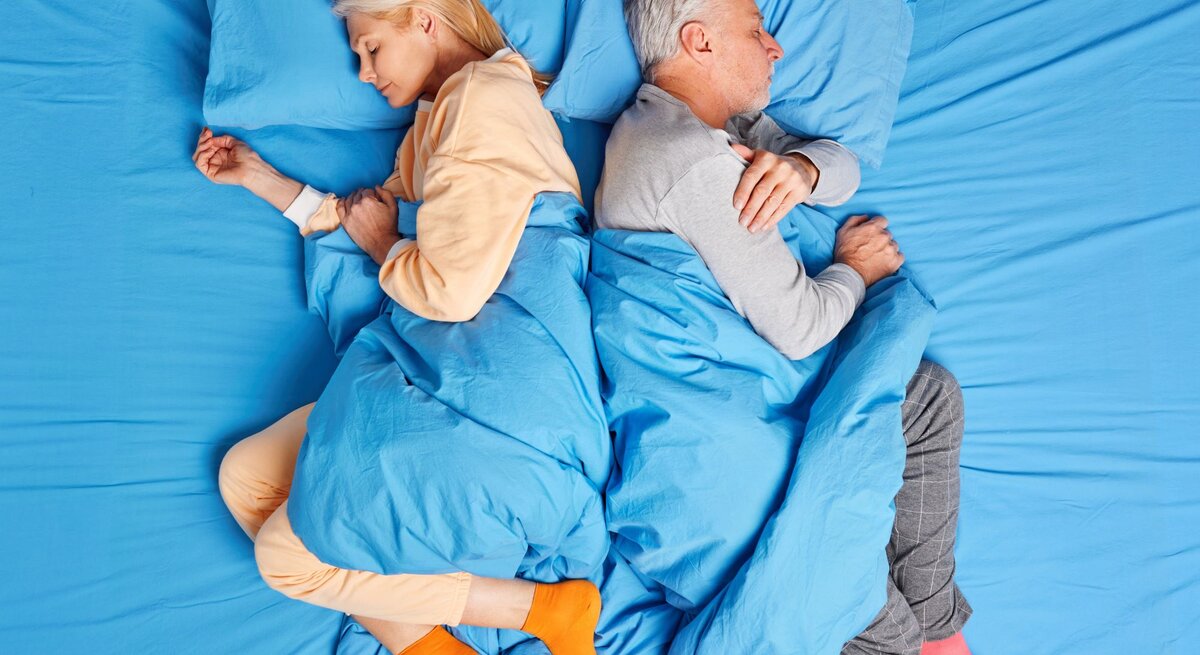 Eine blonde Frau und ein grauhaariger Mann schlafen Rücken an Rücken in farbigen Pyjamas auf blau bezogenem Bett.