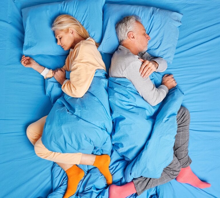 Eine blonde Frau und ein grauhaariger Mann schlafen Rücken an Rücken in farbigen Pyjamas auf blau bezogenem Bett. | © Adobe Stock / wayhome.studio