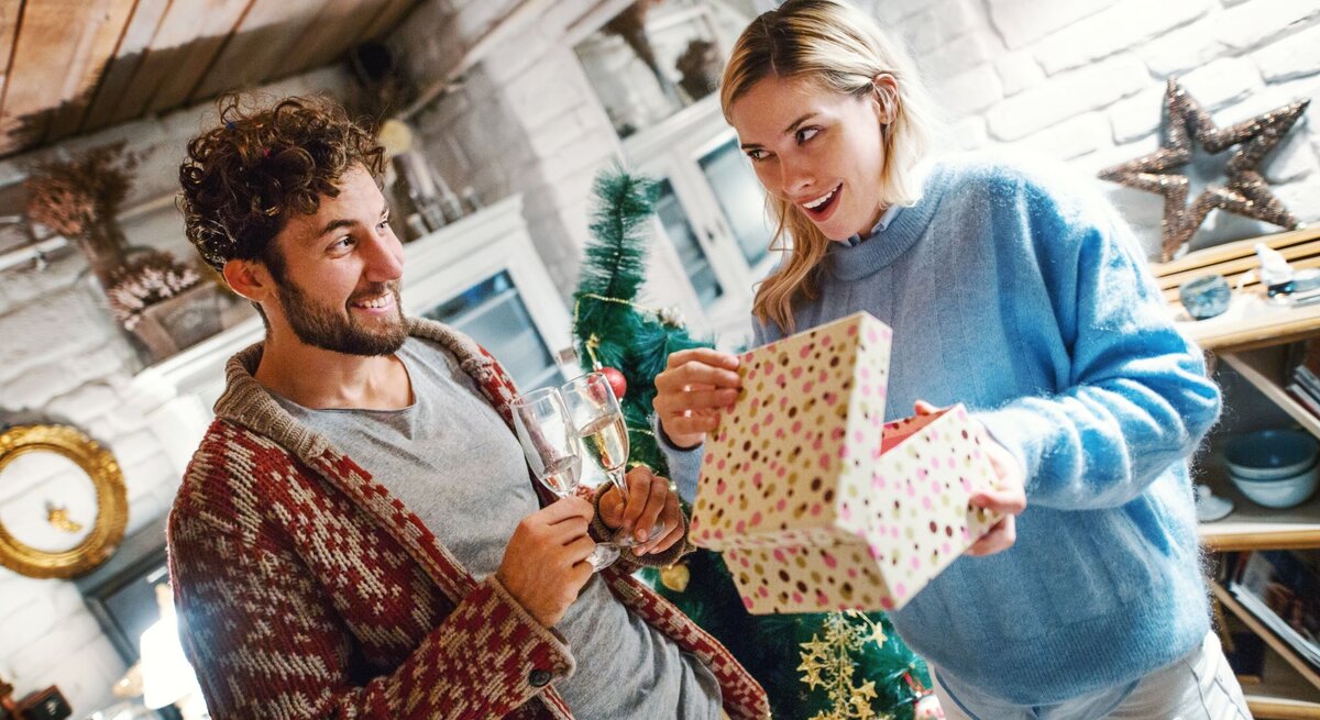 Ein junger Mann mit lockigem Haar und rot gemusterter Strickjacke hält zwei Gläser Sekt, während eine blonde Frau im blauen Pullover lächelnd eine Geschenkbox öffnet.