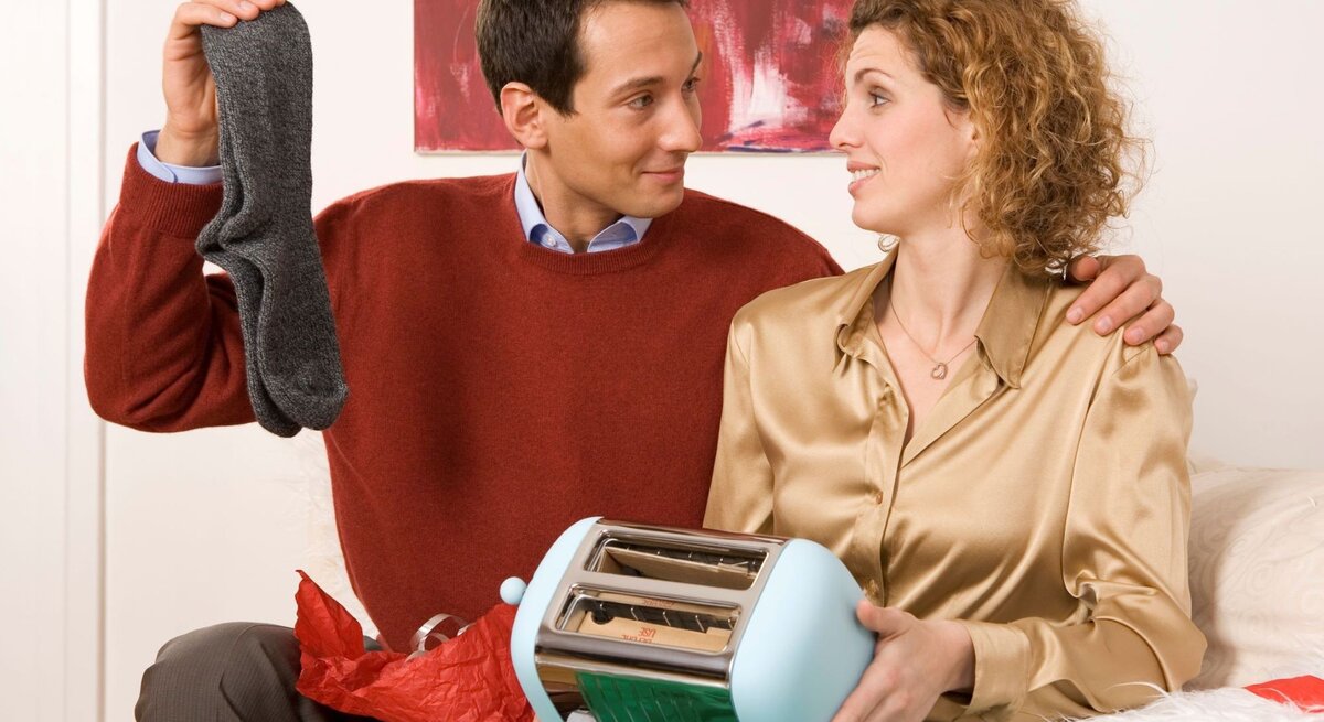 Ein Mann in rotem Pullover hält ein graues Paar Socken hoch, während eine Frau in goldener Bluse mit einem neuen Toaster skeptisch lächelt.