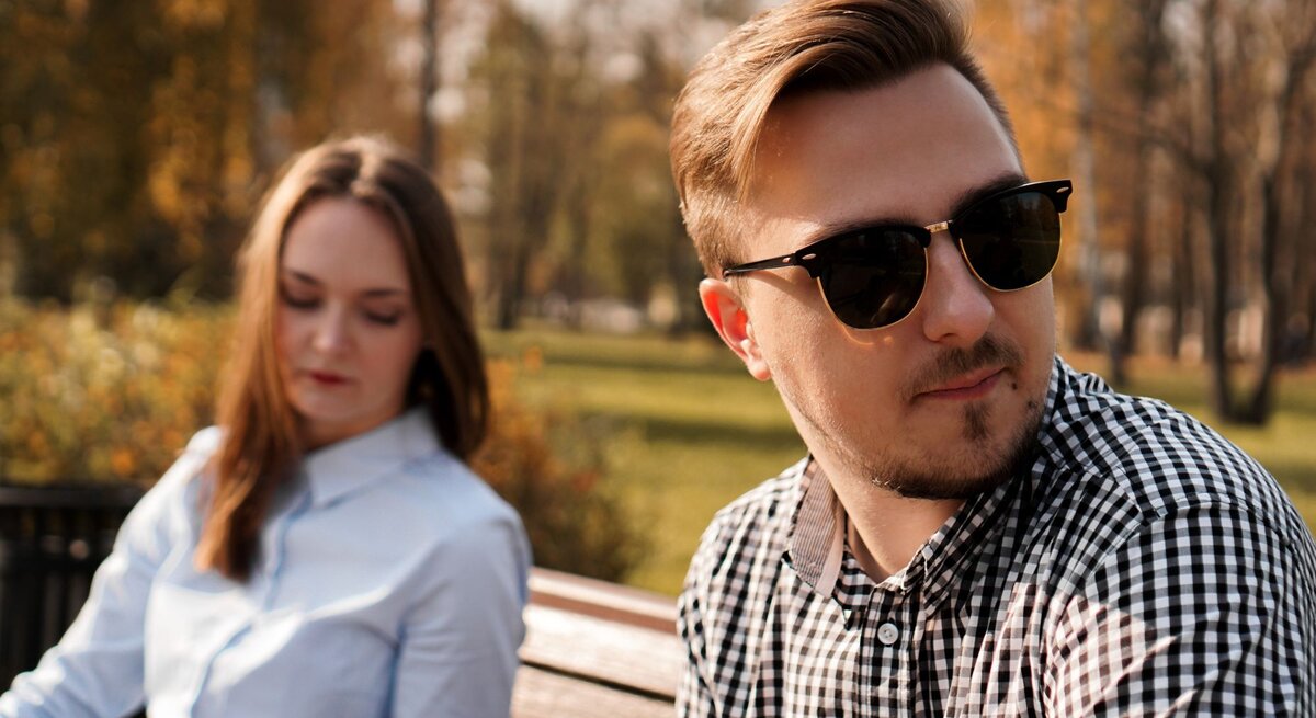 Junges Paar, das auf einer Parkbank im Freien sitzt. Der Mann trägt eine Sonnenbrille, schaut in eine andere Richtung und scheint gedankenverloren zu sein. Die Frau, die neben ihm sitzt, blickt nach unten und wirkt nachdenklich.