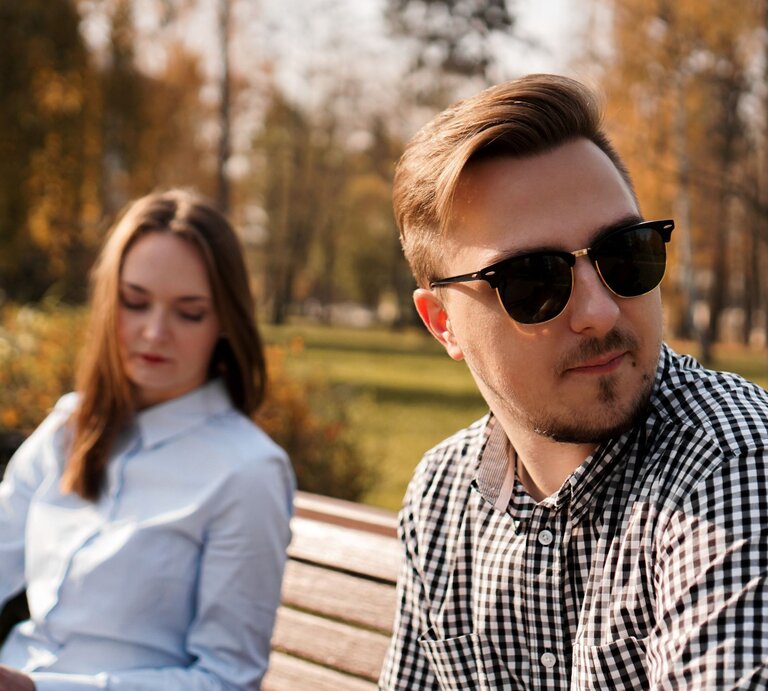 Junges Paar, das auf einer Parkbank im Freien sitzt. Der Mann trägt eine Sonnenbrille, schaut in eine andere Richtung und scheint gedankenverloren zu sein. Die Frau, die neben ihm sitzt, blickt nach unten und wirkt nachdenklich. | © Adobe Stock / brillianata