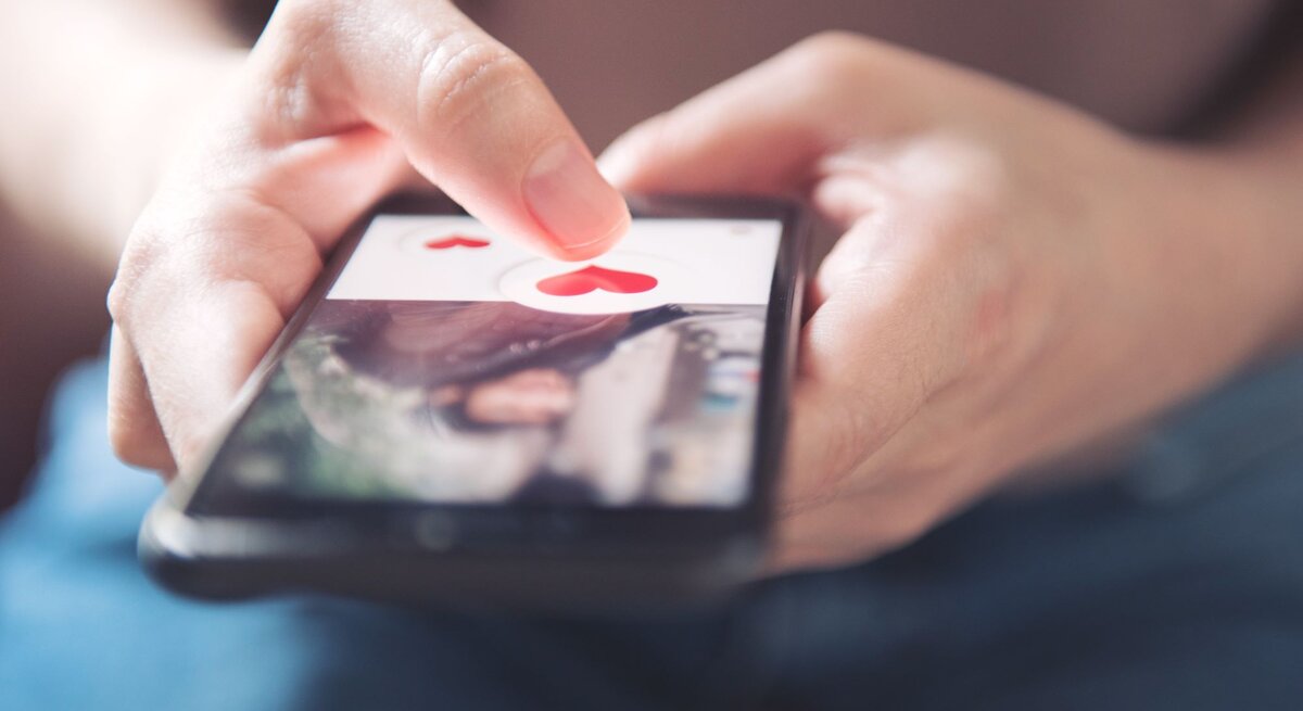Nahaufnahme einer Hand, die ein Smartphone hält. Auf dem Bildschirm ist eine Dating-App geöffnet, die benutzende Person tippt auf ein Symbol in Form eines roten Herzens.