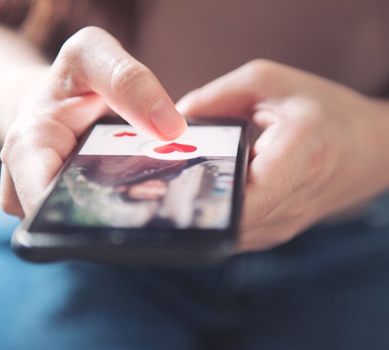 Nahaufnahme einer Hand, die ein Smartphone hält. Auf dem Bildschirm ist eine Dating-App geöffnet, die benutzende Person tippt auf ein Symbol in Form eines roten Herzens. | © Adobe Stock / oatawa