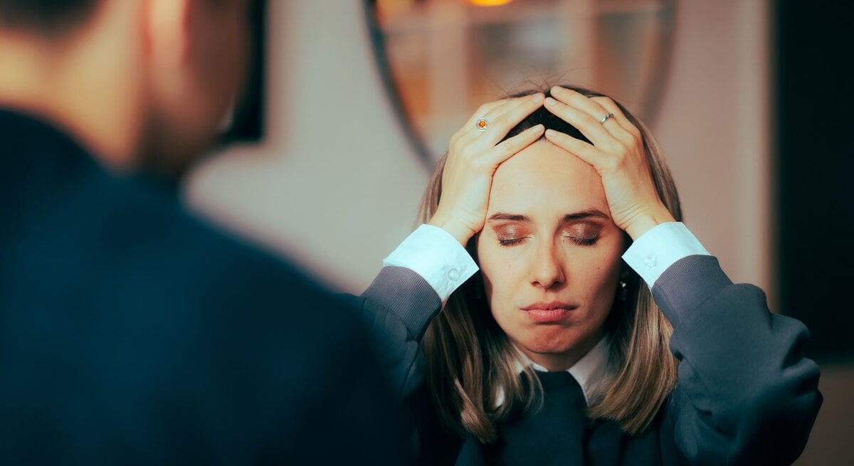 Frau und Mann in einer ernsten oder stressvollen Unterhaltung. Die Frau hält sich mit beiden Händen an den Kopf, die Augen geschlossen und einen besorgten Ausdruck im Gesicht. Der Mann, nur von hinten zu sehen, steht ihr gegenüber.