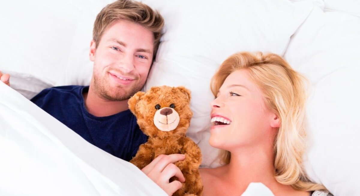 Paar im Bett, das sichtlich Freude hat. Der Mann hält einen braunen Teddybären und blickt in die Kamera. Die blonde Frau neben ihm lacht.