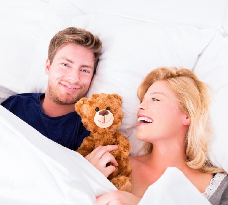 Paar im Bett, das sichtlich Freude hat. Der Mann hält einen braunen Teddybären und blickt in die Kamera. Die blonde Frau neben ihm lacht. | © iStock.com/fizkes