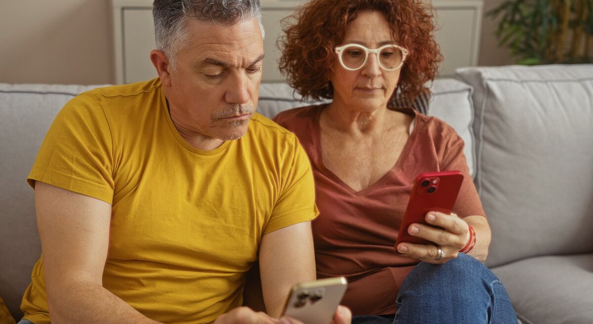 Ein mittelaltes Paar mit Smartphones sitzt konzentriert auf einem grauen Sofa. Der Mann trägt ein senfgelbes Shirt, die Frau eine Brille und rostrote Bluse.