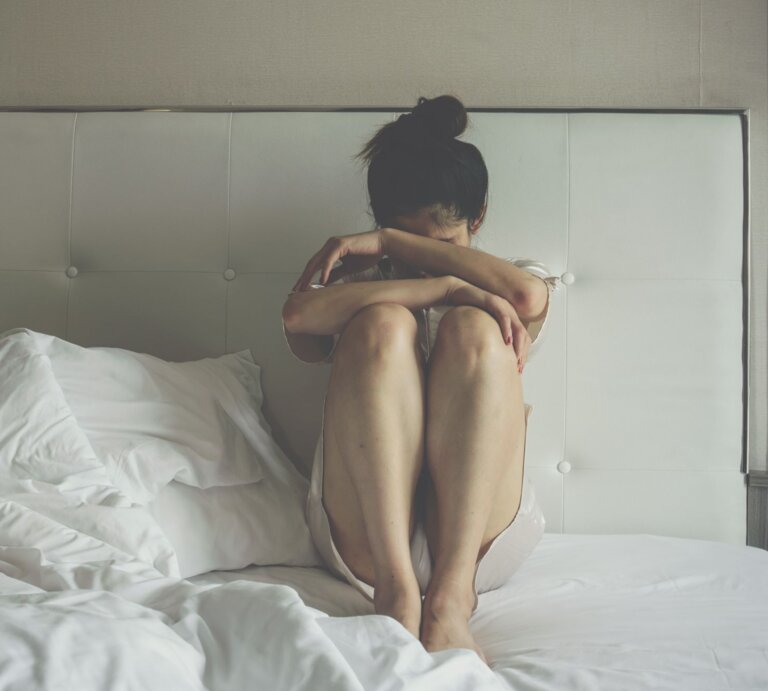 Junge Frau in hellem Nachthemd sitzt verzweifelt mit angezogenen Knien auf einem ungemachten Bett. | © Adobe Stock/ boyloso
