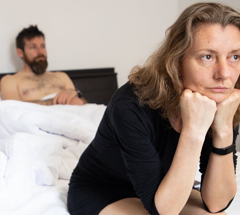 Eine Frau sitzt mit nachdenklicher Haltung im schwarzen Kleid auf einem weißen Bett, im Hintergrund ein Mann auf der Matratze. | © Adobe Stock/ Melinda Nagy