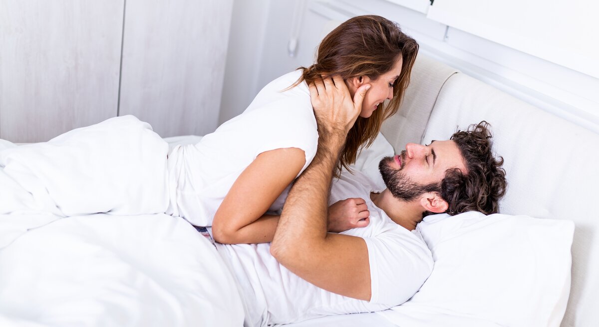 Eine Frau mit braunem Haar kniet auf einem weißen Bett und beugt sich zu einem Mann in weißem Shirt, der auf dem Rücken liegt.