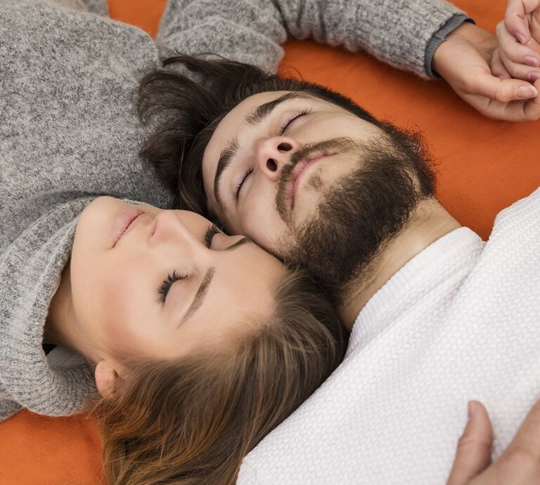 Kuschelndes Paar, das auf einer orangefarbenen Unterlage liegt. Die beiden sind eng umschlungen, ihre Köpfe berühren sich, und sie halten Händchen. | © imago/Westend61