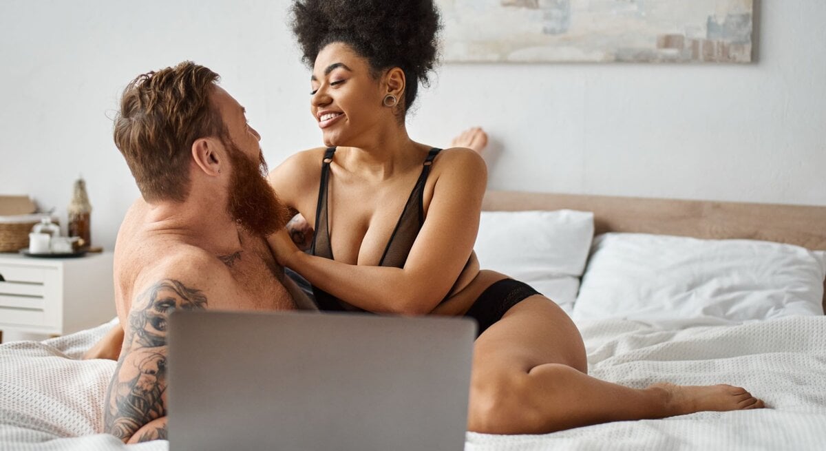 Das Bild zeigt ein Paar, das entspannt und vertraut miteinander auf einem Bett sitzt. Sie lächeln sich an und scheinen einen glücklichen Moment zu teilen. Sie tragen beide legerer Unterwäsche. Auf dem Bett steht ein Laptop, was darauf hindeuten könnte, dass sie gerade gemeinsam einen Porno angesehen haben. Das Zimmer wirkt hell und einladend mit einem großen Kunstwerk an der Wand.