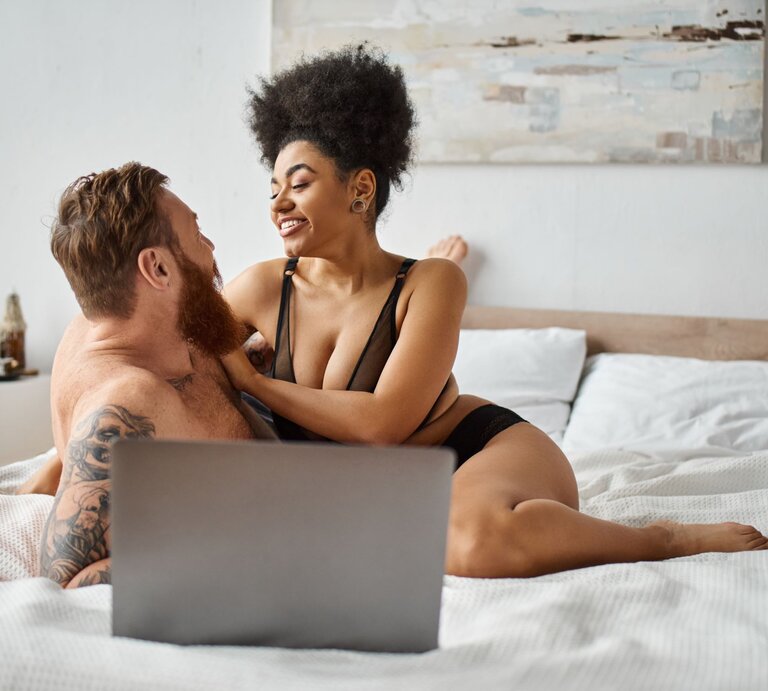 Das Bild zeigt ein Paar, das entspannt und vertraut miteinander auf einem Bett sitzt. Sie lächeln sich an und scheinen einen glücklichen Moment zu teilen. Sie tragen beide legerer Unterwäsche. Auf dem Bett steht ein Laptop, was darauf hindeuten könnte, dass sie gerade gemeinsam einen Porno angesehen haben. Das Zimmer wirkt hell und einladend mit einem großen Kunstwerk an der Wand. | © Adobe Stock/ LIGHTFIELD STUDIOS
