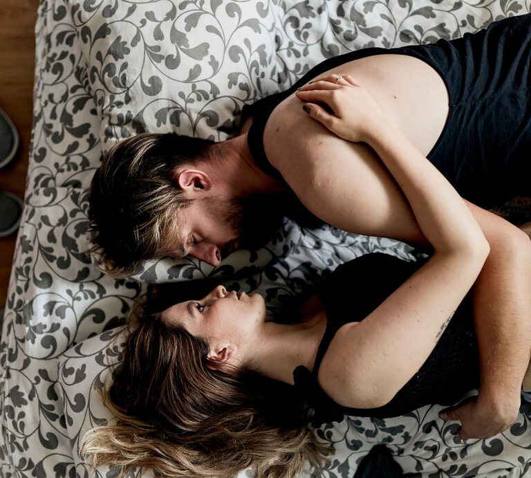Zwei Personen liegen eng umschlungen auf einem Bett mit floralem Muster, Holzfußboden und grauen Hausschuhen im Hintergrund. | © Adobe Stock/ SVP Productions