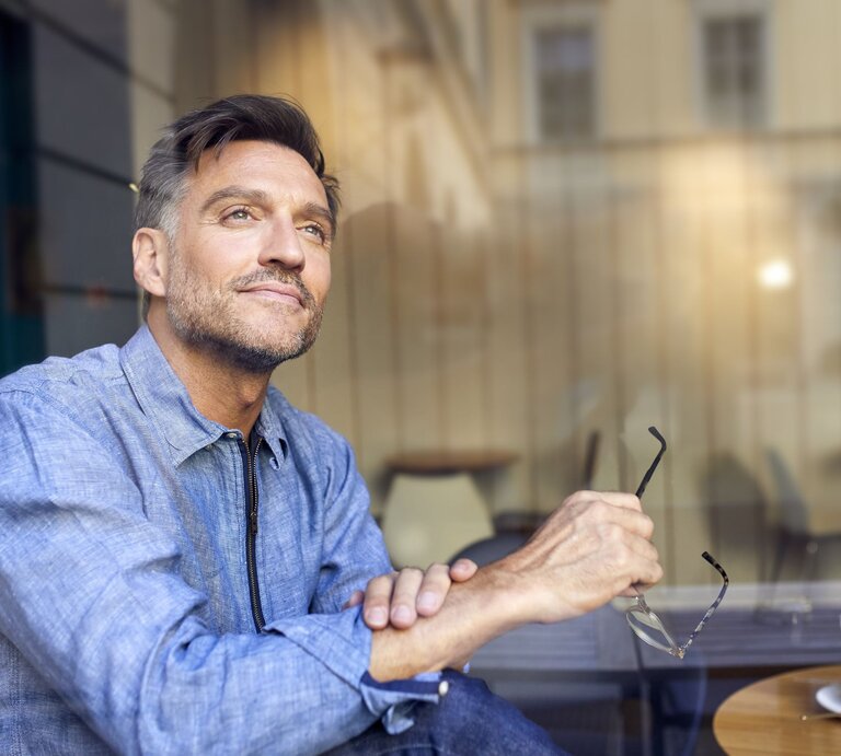 Ein gutaussehender Mann mittleren Alters sitzt an einem Bistrotisch im Freien mit einem Espresso, er schaut verträumt in die Ferne  | © Getty Images /	Westend61