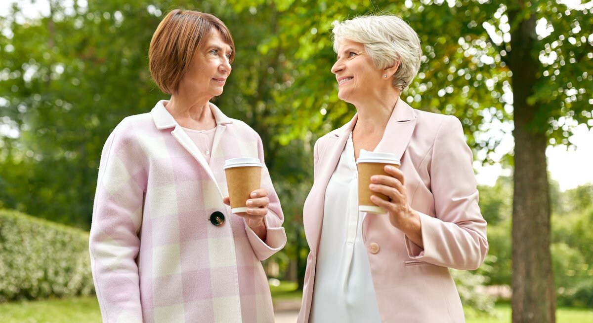 Zwei reife Frauen mit kurzen Frisuren unterhalten sich entspannt mit Coffee-to-go-Bechern in einem sonnigen Park mit Bäumen.