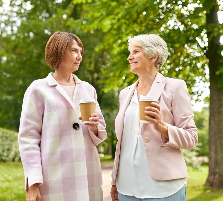 Zwei reife Frauen mit kurzen Frisuren unterhalten sich entspannt mit Coffee-to-go-Bechern in einem sonnigen Park mit Bäumen. | © Adobe Stock / Syda Productions