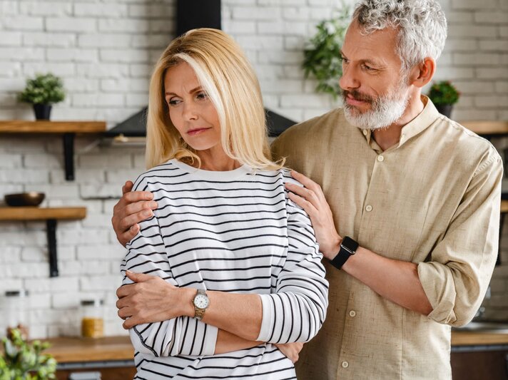 Ein reifer Mann und seine Frau stehen in der Küche, er beruhigt sie und legt ihr beschwichtigend die Hände auf die Schultern. | © Adobe Stock / InsideCreativeHouse
