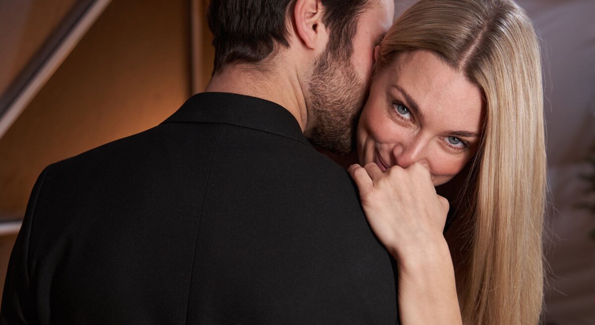 Eine blonde Frau mit langem Haar lächelt verschmitzt, während ein Mann im dunklen Anzug ihr leise etwas ins Ohr flüstert.