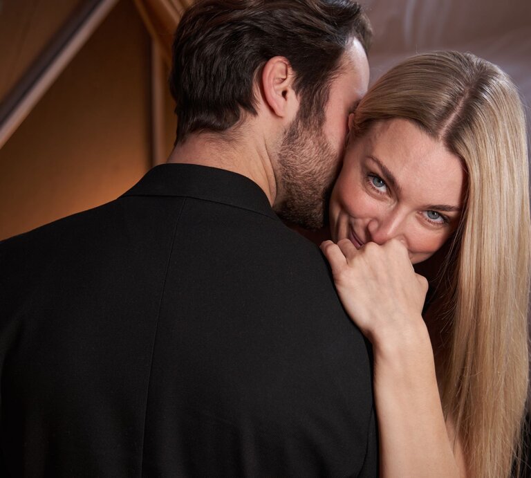 Eine blonde Frau mit langem Haar lächelt verschmitzt, während ein Mann im dunklen Anzug ihr leise etwas ins Ohr flüstert. | © Adobe Stock / Viacheslav Yakobchuk