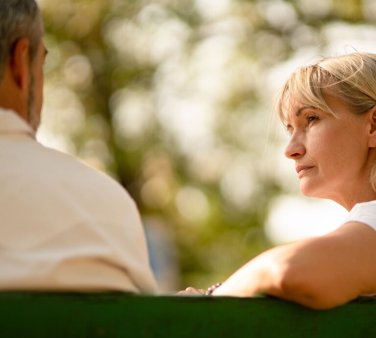 Blonde, nachdenkliche Frau mittleren Alters hört auf einer Parkbank einem Mann mit grauem Haar bei warmem Sonnenlicht zu. | © Adobe Stock / ztony1971