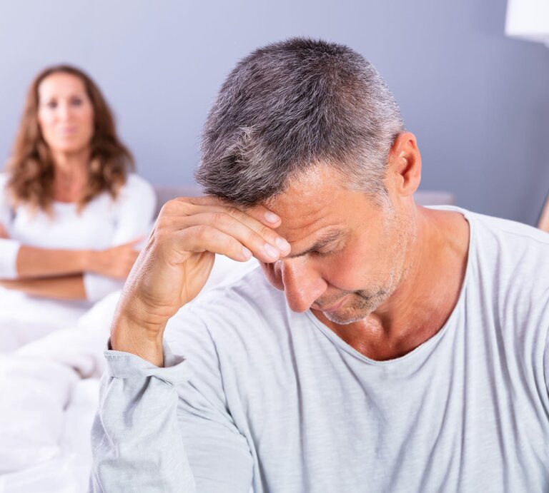 Ein nachdenklicher Mann mit grauem Haar sitzt bedrückt am Bettrand, im Hintergrund blickt eine Frau mit verschränkten Armen ernst. | © Adobe Stock / 