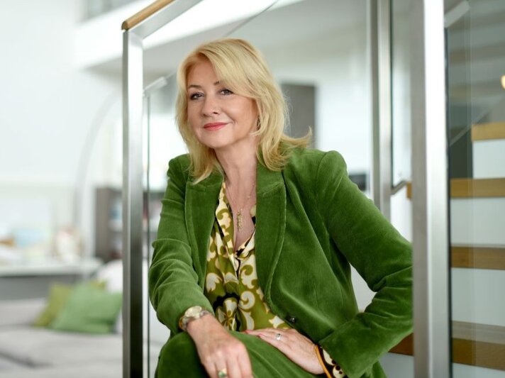 Frau mit blondem Haar trägt grünen Blazer und gemustertes Shirt, sitzt entspannt im modernen Interieur nahe hölzerner Treppe. | © Marzena Seidel