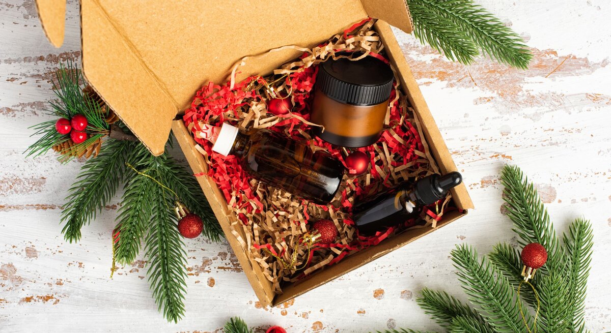 Kosmetik-Set in brauner Box mit rotem Füllmaterial, dekoriert mit Tannengrün und festlichen Christbaumkugeln.