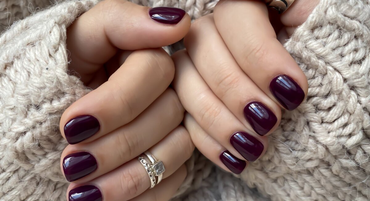 Zwei gepflegte Hände mit dunkelviolett lackierten Nägeln halten sich warm im Ärmel eines beigen Strickpullovers.