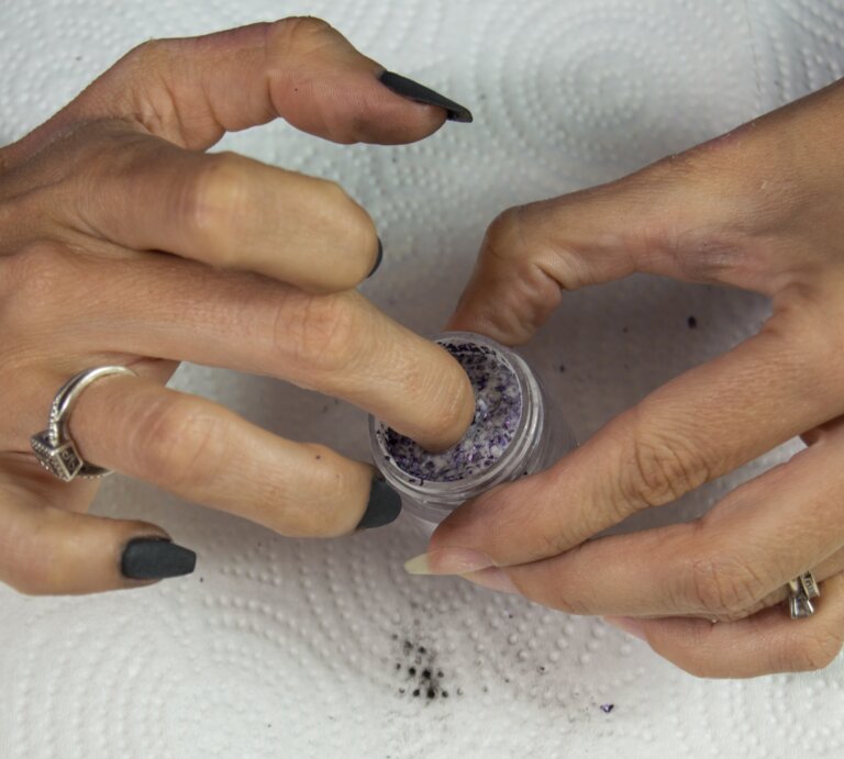 Hände mit dunklem Nagellack, eine Hand taucht Finger in ein kleines Gefäß mit violettem Puder, beide Hände tragen Ringe. | © Shutterstock/ Neal Bryant