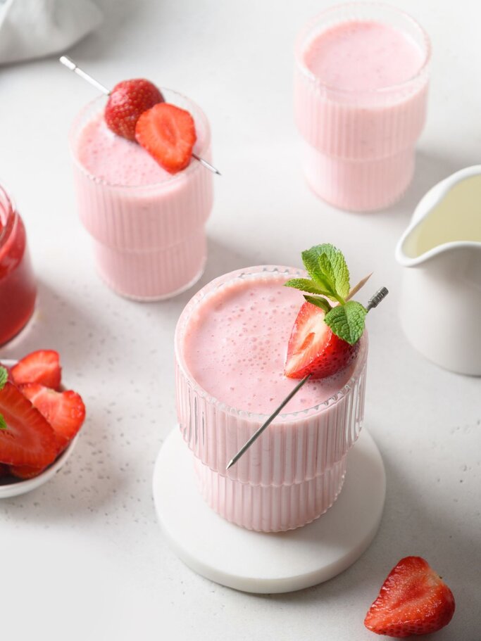 Drei Gläser mit cremigem Erdbeer-Lassi, garniert mit frischen Erdbeeren und Minze, stehen auf hellem Untergrund. | © Getty Images/ Svetlana-Cherruty