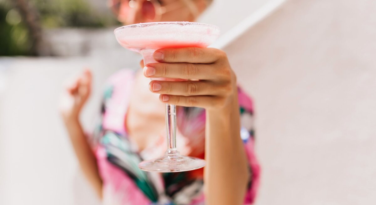 Verschwommenes Porträt einer Frau mit Sonnenbrille, die einen rosa Cocktail in die Kamera hält, vor heller Kulisse.