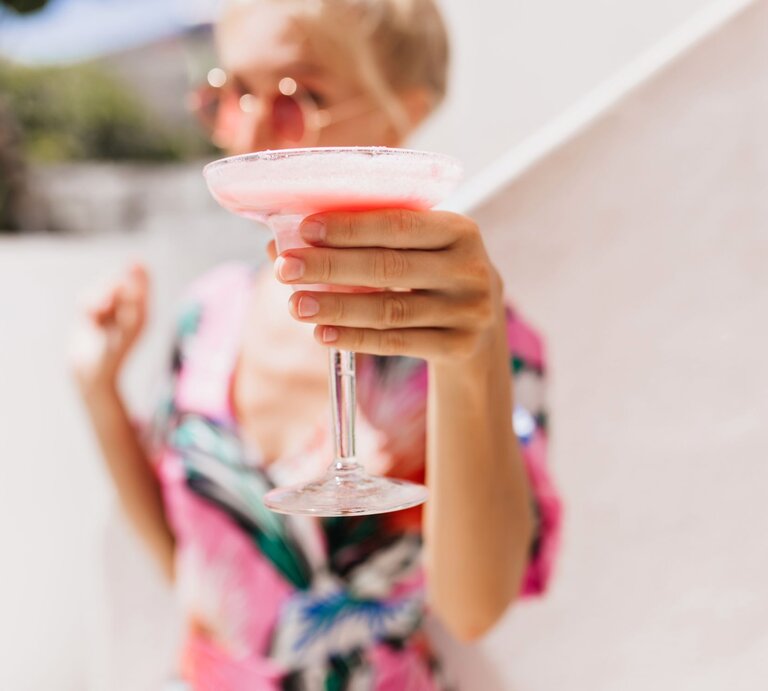 Verschwommenes Porträt einer Frau mit Sonnenbrille, die einen rosa Cocktail in die Kamera hält, vor heller Kulisse. | © Glow25
