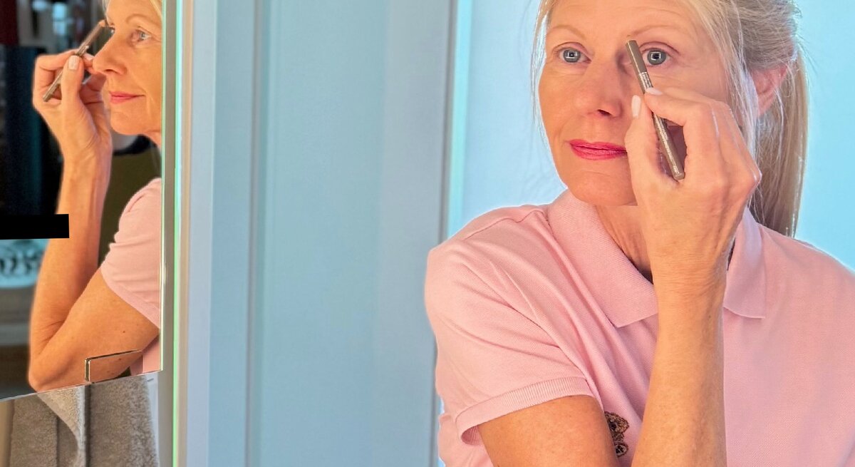 Blonde Renate Zott im mittleren Alters trägt konzentriert Augen-Make-up auf, ihr Spiegelbild ist links im hellen Badezimmer zu sehen.