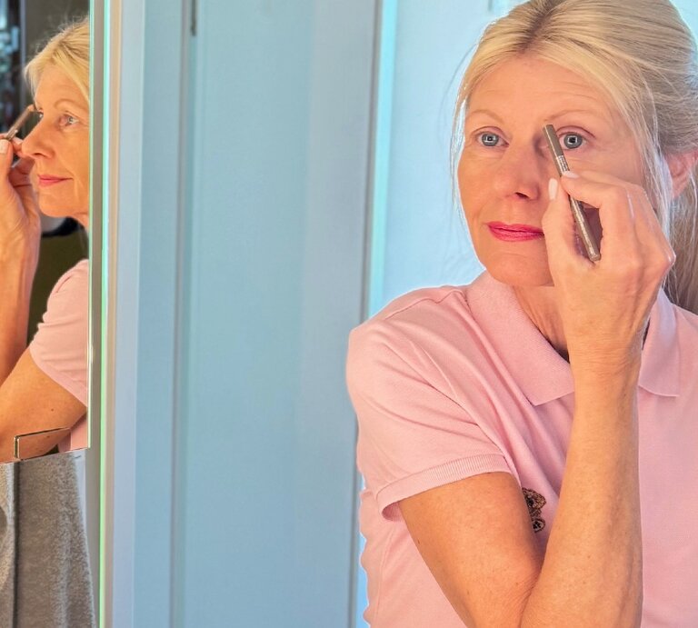 Blonde Renate Zott im mittleren Alters trägt konzentriert Augen-Make-up auf, ihr Spiegelbild ist links im hellen Badezimmer zu sehen. | © Renate Zott