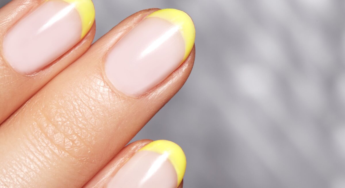 Nahaufnahme einer gepflegten Hand mit pastellrosa Nägeln und leuchtend gelben Spitzen vor einem verschwommenen grauen Hintergrund.