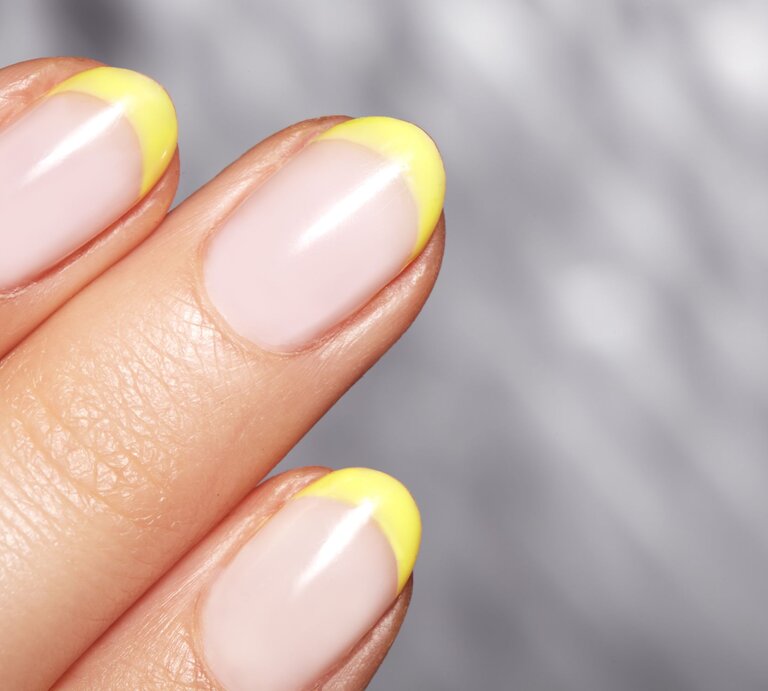 Nahaufnahme einer gepflegten Hand mit pastellrosa Nägeln und leuchtend gelben Spitzen vor einem verschwommenen grauen Hintergrund. | © Adobe Stock/ marinafrost
