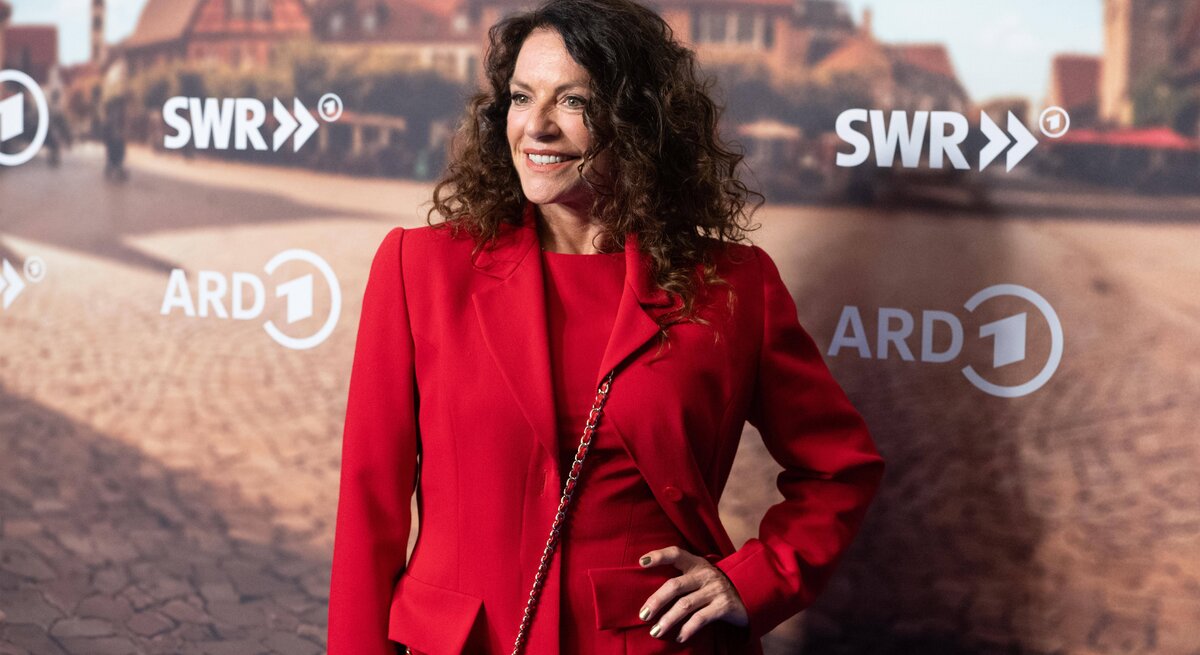 Christine Neubauer mit dunklen lockigen Haaren trägt ein elegantes rotes Outfit, dazu goldenen Nagellack und eine passende rote Tasche vor einem städtischen Hintergrund.